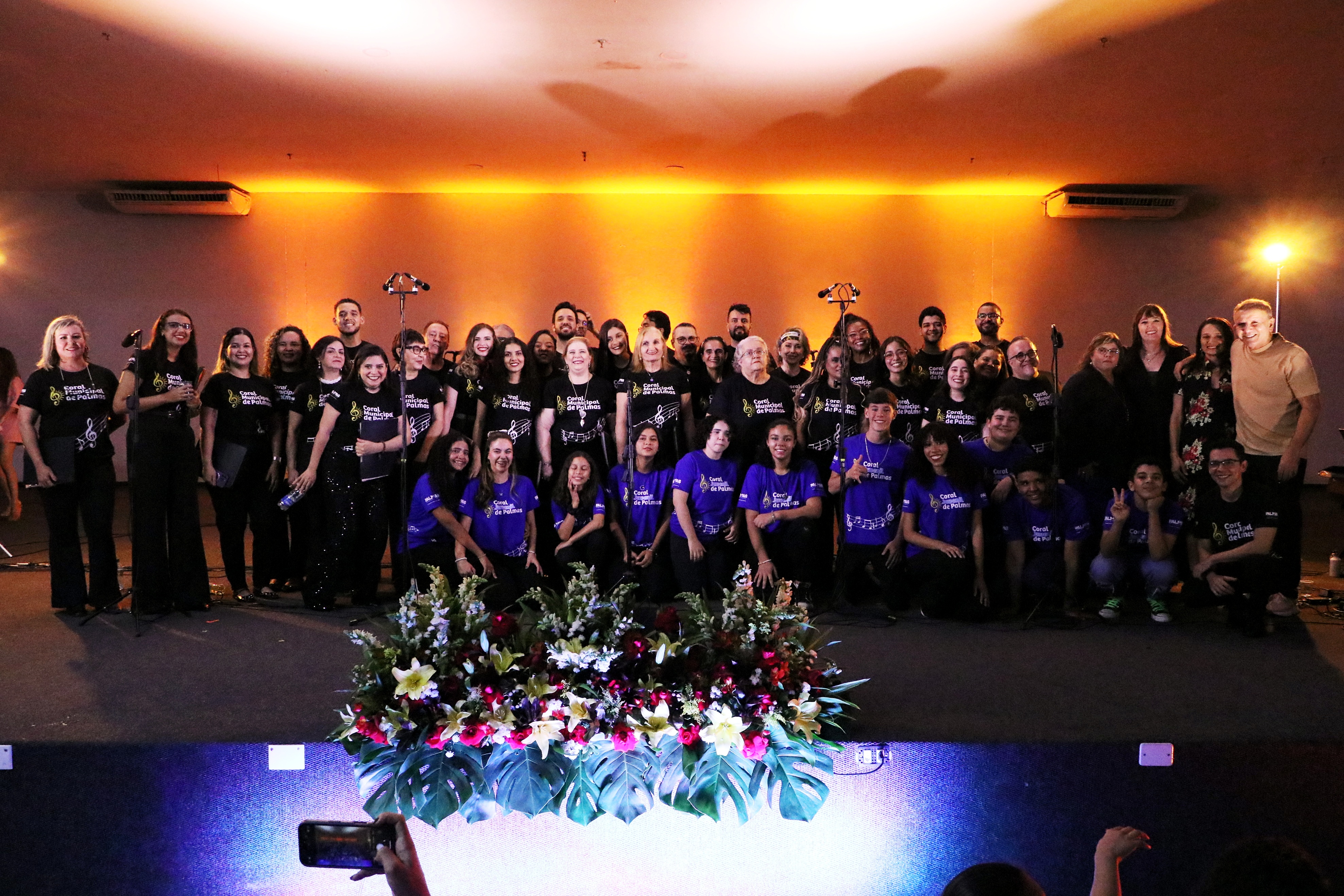 O prefeito Eduardo (à direita, de pé), a primeira-dama Polyanna, a maestrina Renate e a presidente Luara posam junto aos corais Municipal e Juvenil de Palmas após o encerramento do ‘Concerto Rock.Br’